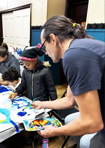 Caring male art teacher encouraging a young student during a creative painting project at The Belfry Centre in Brooklyn.